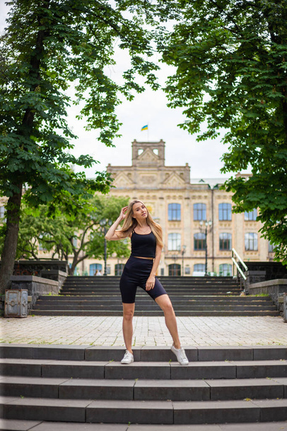 Young beautiful blonde woman with  athletic build in a black t-shirt and in black tight sports shorts stands on the stairs and looks straight - Valokuva, kuva