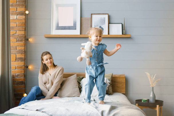 Een schattig twee-jarig meisje in een spijkerbroek met een zacht speeltje in haar handen springt op het bed, en haar moeder zit en kijkt naar haar - Foto, afbeelding