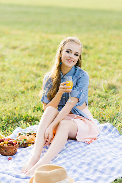 Una bella ragazza con un bicchiere di succo appena spremuto siede su una coperta a un picnic estivo e sorride un sorriso affascinante - Foto, immagini