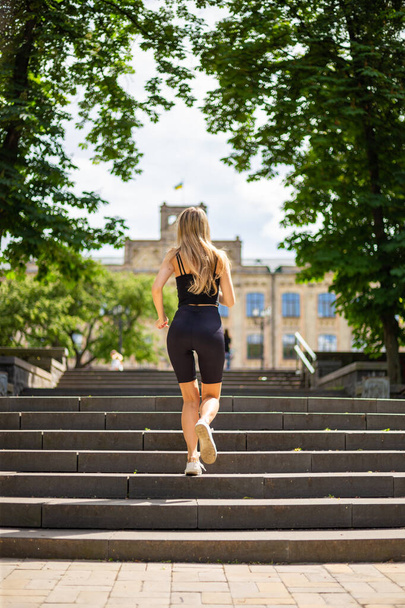 Une jeune belle femme blonde au physique sportif en T-shirt noir et en short de sport noir serré monte les escaliers - Photo, image