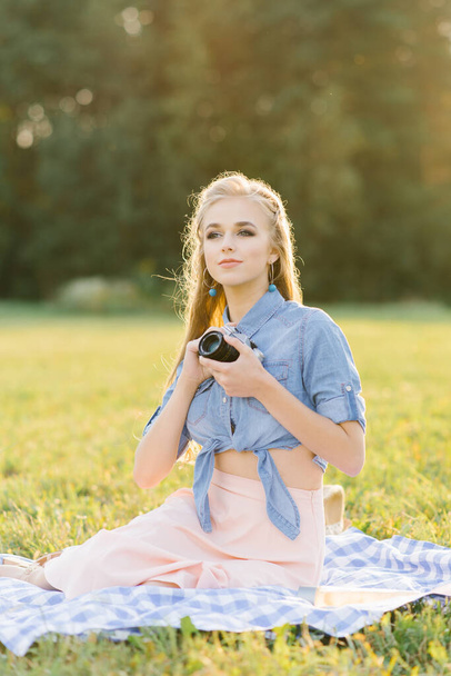 Μια νεαρή όμορφη γυναίκα κάθεται σε μια κουβέρτα στη φύση και κρατά μια κάμερα στα χέρια της - Φωτογραφία, εικόνα