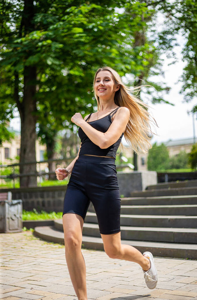 Uma jovem mulher loira bonita com um físico esportivo em uma camiseta preta e em shorts de esportes apertados pretos sobe as escadas - Foto, Imagem