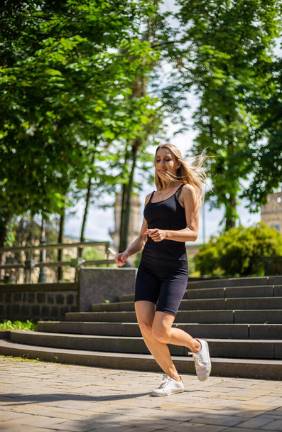 Une jeune belle femme blonde au physique sportif en T-shirt noir et en short de sport noir serré monte les escaliers - Photo, image