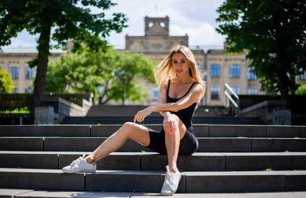 Young beautiful blonde sportive woman in a black tank top and in black tight sports shorts sits on the stairs - Foto, immagini