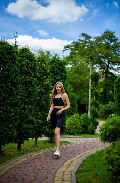 Young beautiful sporty blonde woman in a black T-shirt and in black tight sports shorts in good shape posing in front of green trees and bushes in nature - 写真・画像
