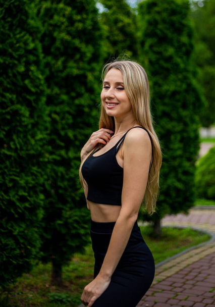 Young beautiful sporty blonde woman in a black T-shirt and in black tight sports shorts in good shape posing in front of green trees and bushes in nature - Foto, imagen