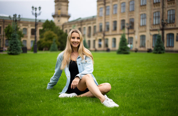 Young beautiful modern fashionable girl in a denim jacket sits on a green grass - Foto, imagen