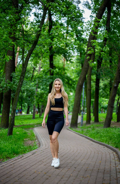 Fiatal, gyönyörű, sportos szőke nő fekete pólóban és fekete szűk sportnadrágban, jó formában, zöld fák és bokrok előtt pózolva a természetben. - Fotó, kép