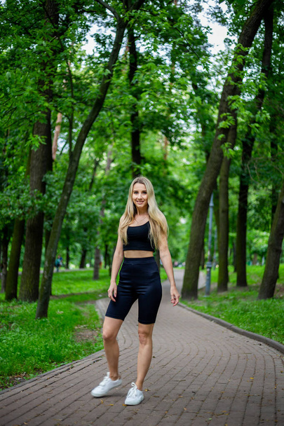 Young beautiful sporty blonde woman in a black T-shirt and in black tight sports shorts in good shape posing in front of green trees and bushes in nature - Foto, Bild