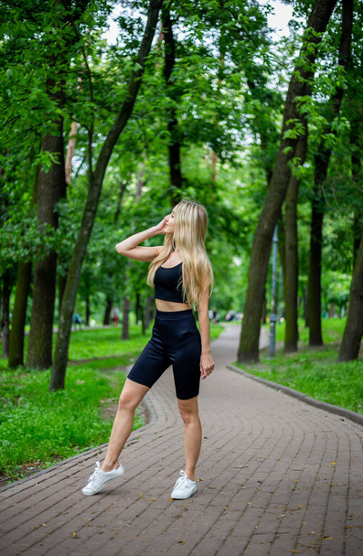 Молода красива спортивна блондинка в чорній футболці і в чорних жорстких спортивних шортах в хорошій формі позує перед зеленими деревами і кущами на природі
 - Фото, зображення