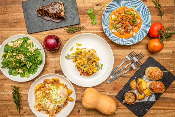 Blick von oben auf köstliche mediterrane Gerichte auf einem Holztisch. Gebratene Nudeln, Spinat und Pinienkernsalat, Spiegeleier mit Kartoffeln. Kürbiscreme, grüne Tomaten - Foto, Bild