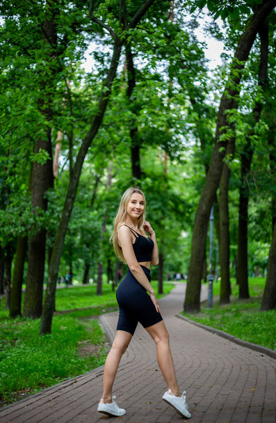 Young beautiful sporty blonde woman in a black T-shirt and in black tight sports shorts in good shape posing in front of green trees and bushes in nature - Zdjęcie, obraz