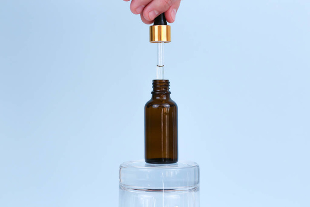 Male hand holds dropper with oil. Opened jar of natural cosmetic product on glass podium on blue background. Essential oil for care of skin. Skincare, moisturizing beauty product. Cosmetic production. - Photo, Image