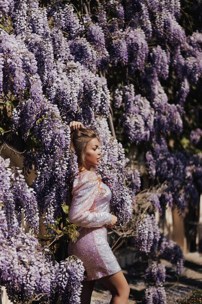 fashion outdoor photo of beautiful woman with blond hair in elegant dress and accessories posing near blooming wisteria tree - Foto, Imagem