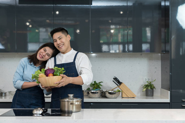 Retrato de una encantadora pareja en un delantal sosteniendo una cesta de verduras para cocinar alimentos en una cocina casera. Pareja asiática utiliza el tiempo libre para cocinar juntos los fines de semana en la isla de la cocina. Estilo de vida culinario - Foto, Imagen
