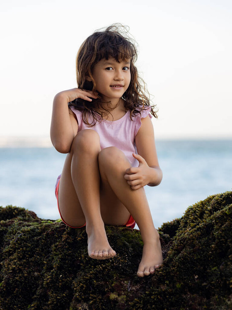 Молода дівчина сидить на скелі біля океану і посміхається. Щасливого дитинства. Проводжу час на пляжі. Мила дівчинка. Відпустка в Азії. Літня концепція. Пляж Пандава, Балі, Індонезія - Фото, зображення