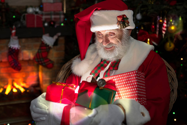 Άγιος Βασίλης με δώρο κουτί παρουσιάζει κοντά στο τζάκι και χριστουγεννιάτικο δέντρο. Εορταστική εσωτερικό ξύλινο σπίτι, χαρούμενη διάθεση Πρωτοχρονιάς Πνεύμα των Χριστουγέννων. Ανώτερος άνθρωπος με πραγματική λευκή γενειάδα cosplay Άγιος Βασίλης. - Φωτογραφία, εικόνα