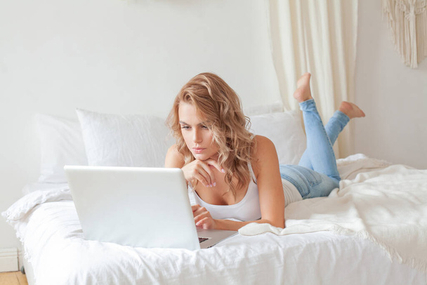 Young woman in casual clothes with her legs up liying on sofa at cozy home interior and using laptop - Фото, зображення