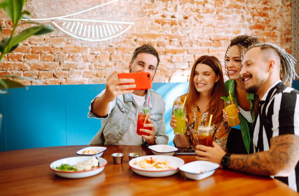 Selfie time! Young friends take a selfie and smile while relaxing in a cafe. Two women and two men in a cafe talking, laughing, drinking cocktails and eating. People, lifestyle, lunch, party. - Foto, Imagem