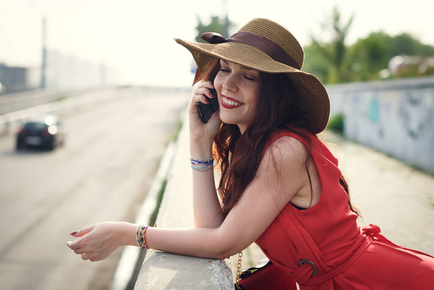 A happy young woman in a wide-brimmed hat is walking in the summer with a bag and talking on the phone - Foto, imagen