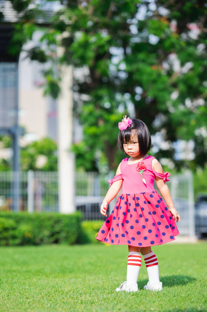 Asian girl stands frowning on grass after her losing game. Sportsmanship. Children are not satisfied. Child do not accept defeat. Kid wears pink dress. Female wear long white socks. Baby 4-5 years old - Foto, afbeelding