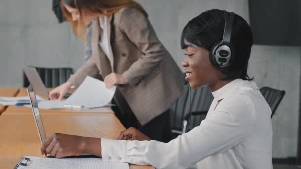 Young african business woman working call center manager support line agent speaks remote communication wears headset headphones advises client makes videocall in office on background of colleagues - Footage, Video