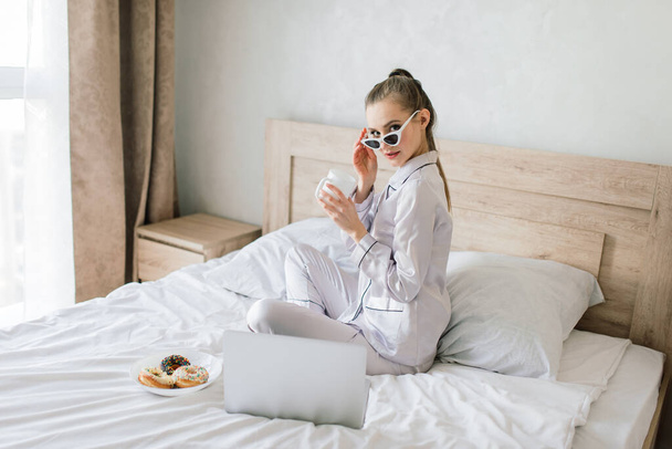 Foto de una joven mujer alegre en pijama escribiendo en el portátil y sonriendo mientras está sentada en la cama en la sala de luz - Foto, imagen