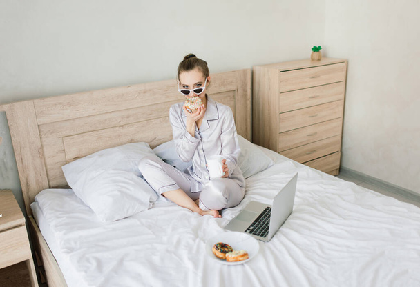 Foto de una joven mujer alegre en pijama escribiendo en el portátil y sonriendo mientras está sentada en la cama en la sala de luz - Foto, imagen