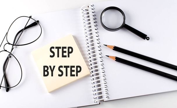 Слово STEP BY STEP на хворий на блокноті з олівцями, магнітним підсилювачем та окулярами
 - Фото, зображення