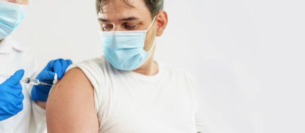 doctor gives an adult young man in a medical mask an injection in the shoulder. Vaccination of adults against coronavirus. The vaccine against Covid-19. Protection against the virus. Copy spase - Photo, image