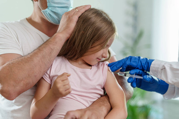 doctor gives a brave little girl an injection in the shoulder. the child sits on parent's lap, is not afraid and smiles.Vaccination of children against coronavirus. vaccine against Covid-19. Close-up - Photo, image
