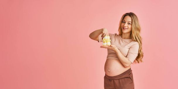 attractive young pregnant woman, holding socks for a newborn near her stomach, standing on a pink background and looking down. Happy pregnancy, waiting for the baby. Copy space - Foto, immagini
