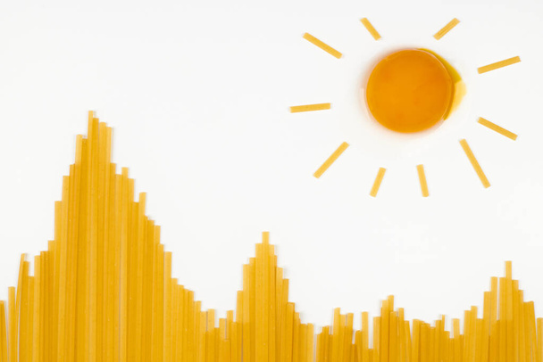 Sonne in den Bergen. Ungekochte Spaghetti in Form von Hügeln und Ei als Sonne. Reisesnack-Konzept. Vereinzelter weißer Hintergrund. Ein Bündel italienische Pasta von oben, flach gelegt. - Foto, Bild