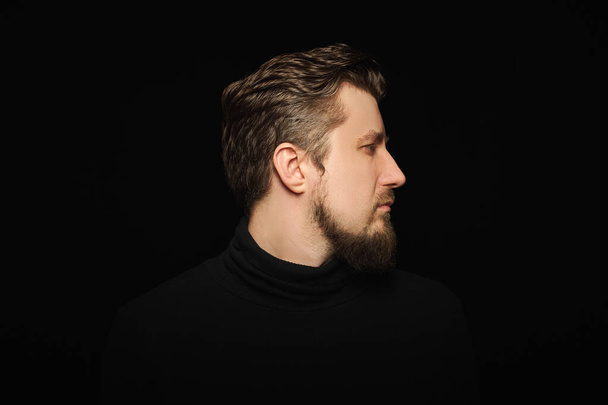 Thoughtful bearded man, half profile portrait, black background. Young guy in black polo neck sweater. Reflecting and thinking successful man. - Фото, изображение
