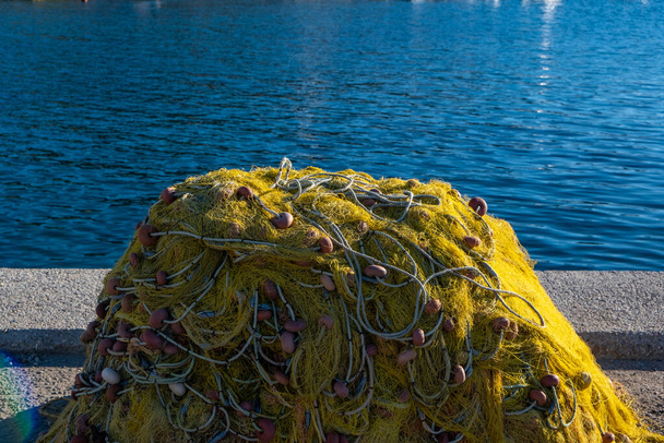 Τα δίχτυα αλιείας στοιβάζονται στην αποβάθρα του λιμανιού και στεγνώνουν κάτω από τον ήλιο. Κίτρινο χρώμα διχτυωτά με σχοινιά και πλωτήρες σε κυματιστό φόντο θάλασσας. Ελληνικό νησί Κυκλάδες εξοπλισμός ψαράδων. - Φωτογραφία, εικόνα