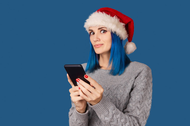 Beautiful happy young woman in santa hat with smartphone over blue background - Photo, Image