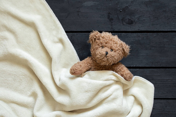 Oyuncak ayı kahverengi ayı beyaz bir battaniyenin altında yatıyor, siyah tahta bir arka planda, çocuk. - Fotoğraf, Görsel