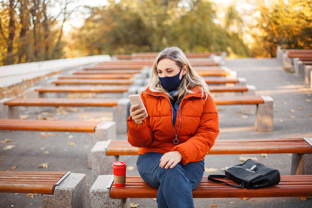 Mulher loira em outerwear e máscara andando no caminho pavimentado no dia ensolarado no parque de outono durante a pandemia - Foto, Imagem