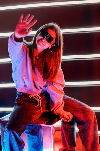 Jonge vrouw in futuristische bril aanraken hoofd en weg te kijken onder kleurrijke neon verlichting - Foto, afbeelding
