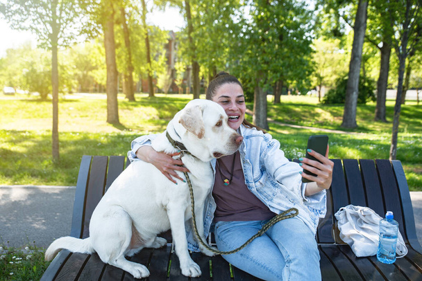 Young beautiful woman sitting with her pet dog a golden Labrador Retriever on a bench wants to take a selfie with a smartphone  - Foto, Imagen