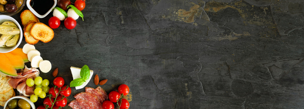 Italian theme charcuterie corner border against a dark slate banner background. Selection of cheese, meat and fruit appetizers. Above view with copy space. - Fotoğraf, Görsel