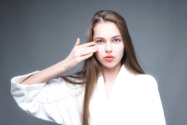 A beautiful girl in a bathrobe applies a mask to her face with her hand. Evening skin care - Φωτογραφία, εικόνα