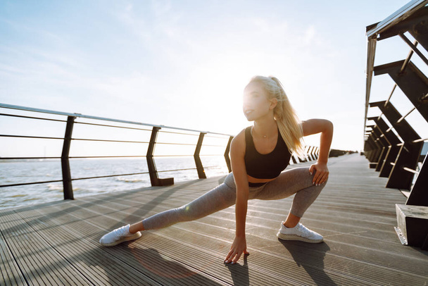 Young fitness woman exercising and stretching outdoors in the morning. Fit healthy athlete is doing workout at the beach pier. Sport, Active life. - Fotó, kép