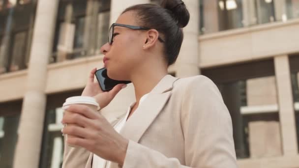 Low-angle tracking slowmo shot of young beautiful businesswoman in smart casual wear and brille talking on phone outdoor in business district then winkt jemandem mit Lächeln - Filmmaterial, Video