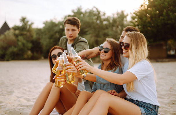 The young friends holding bottles with beer at the beach party. Group of friends having fun, relaxing, toasting with beerus. People, lifestyle, travel, nature and vacations concept. - Фото, изображение