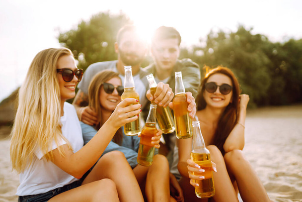 The young friends holding bottles with beer at the beach party. Group of friends having fun, relaxing, toasting with beerus. People, lifestyle, travel, nature and vacations concept. - Foto, afbeelding