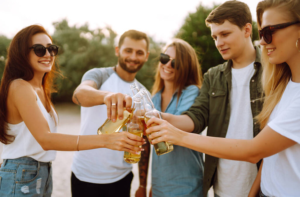 The young friends holding bottles with beer at the beach party. Group of friends having fun, relaxing, toasting with beerus. People, lifestyle, travel, nature and vacations concept. - Φωτογραφία, εικόνα