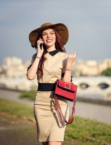 A happy young woman in a wide-brimmed hat is walking in the park in the summer with a bag and talking on the phone - Foto, imagen