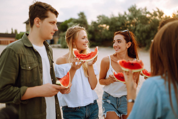 Group of young friends having picnic, eating watermelon, enjoy summer party at the beach. Happy friends resting together sitting near the sea. Beach holiday and summer vacation concept. - Foto, Bild
