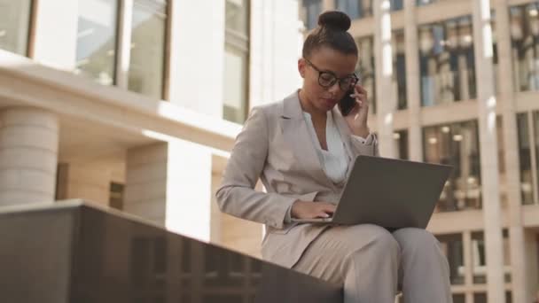 Plan moyen d'une jeune femme d'affaires sérieuse en tenue décontractée intelligente parlant au téléphone avec un partenaire d'affaires ou un investisseur prenant des notes dans un ordinateur portable assis à l'extérieur dans le centre-ville - Séquence, vidéo